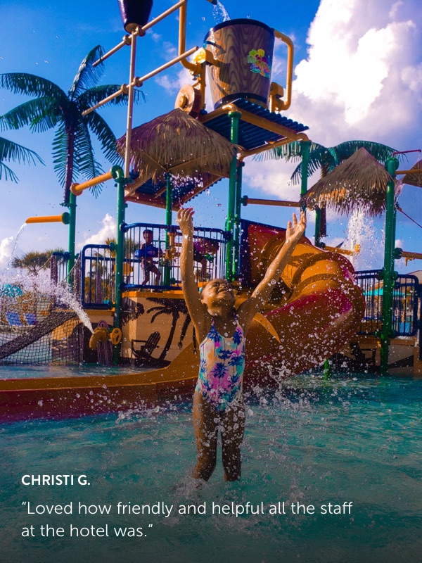 Photo submission from Christi G. showing a small child standing in a kiddie pool at a water park with arms outstretched splashing water.