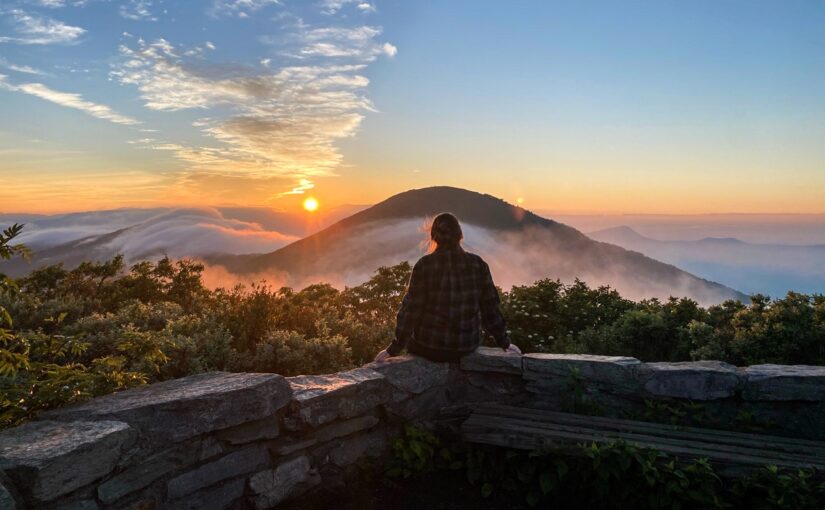 A woman sitting on a stone wall facing a mountain scene during sunrise.
